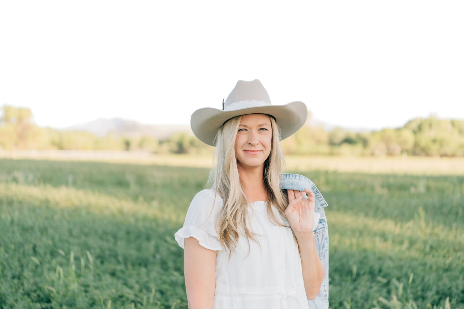 Smiling blonde woman in white dress and tan cowboy hat standing in green field with mountains in background. Peoria Arizona