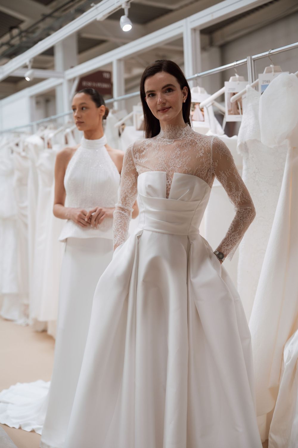 Two models wearing elegant white wedding gowns pose in a bridal boutique surrounded by dresses on racks.