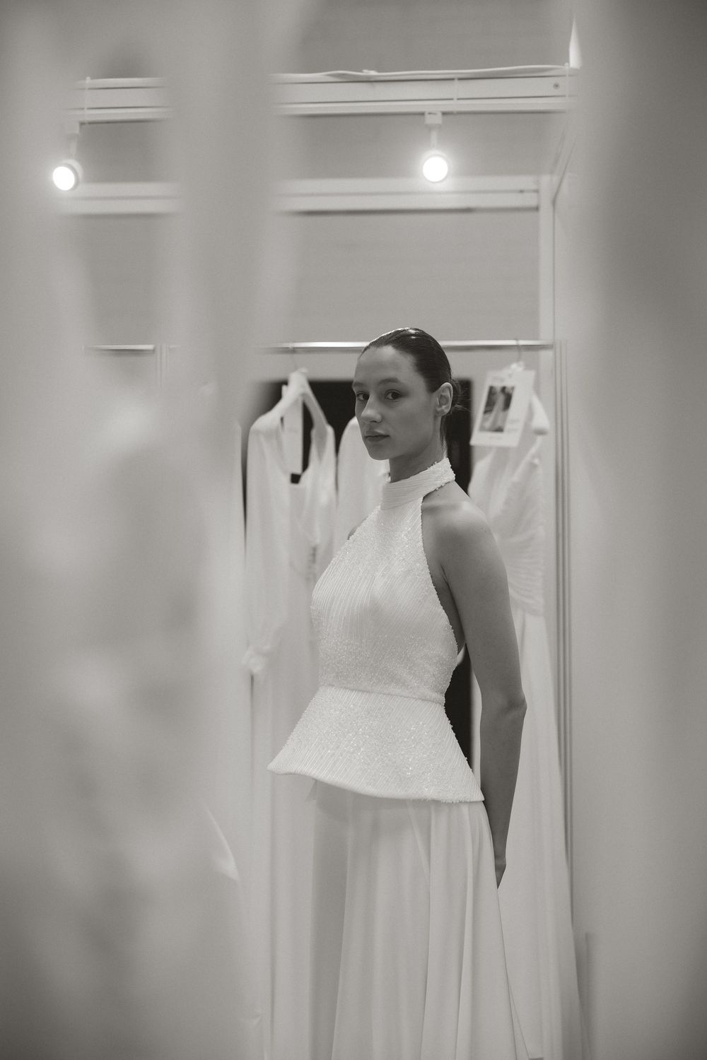 Black and white photo of a woman in a white halter peplum wedding gown posing in a bridal boutique.