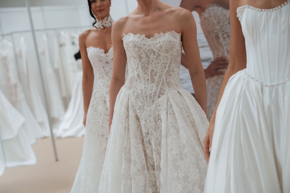 Three women wearing elegant strapless white lace and satin wedding gowns displayed in a bridal boutique.