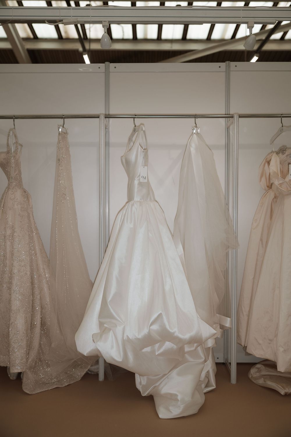 White wedding gowns displayed on hangers in a bridal boutique showroom, featuring elegant satin and lace designs.