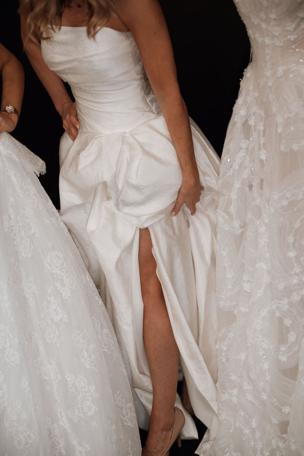 Model wearing a strapless white satin bridal gown with dramatic ruched skirt and thigh-high slit on runway.