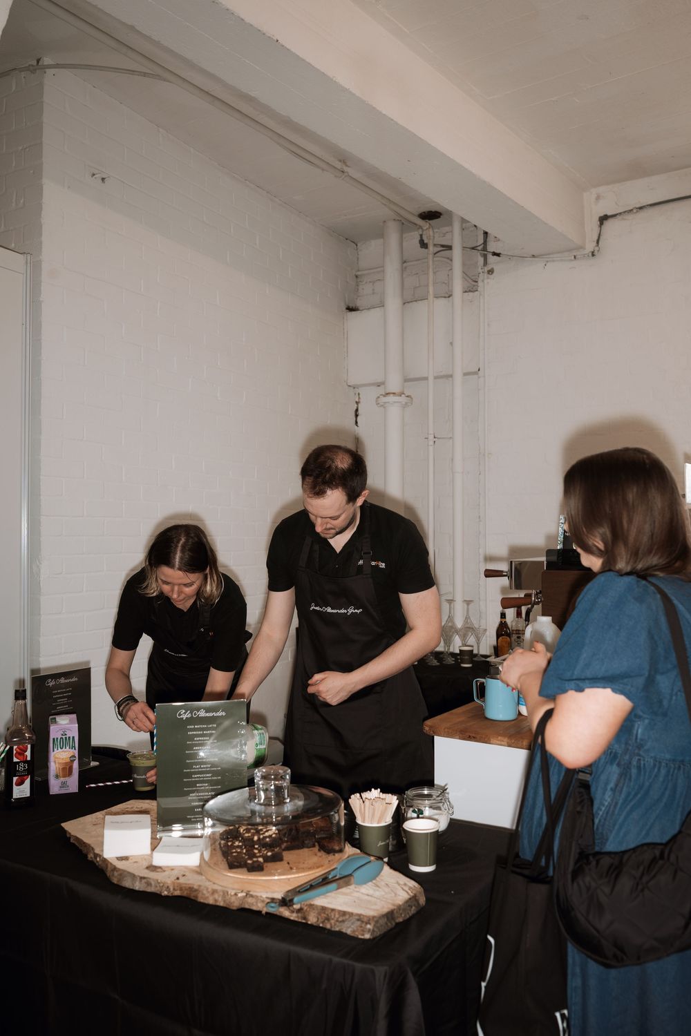 Two baristas in black aprons prepare drinks at a pop-up coffee stand for a customer in a bright indoor venue.