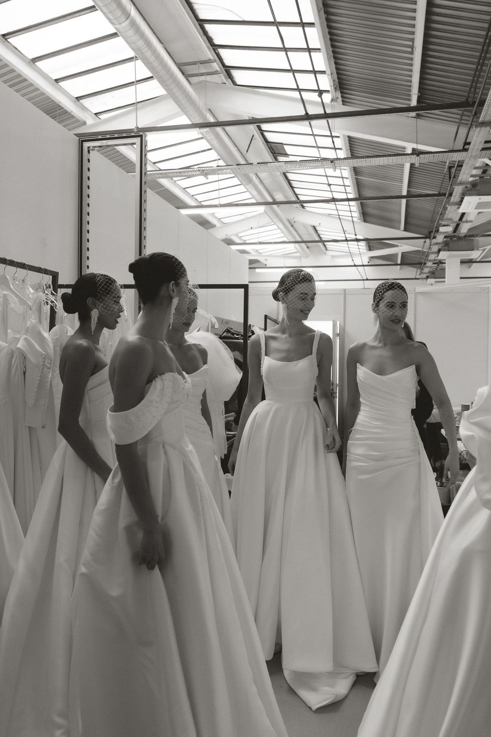 Black and white photo of three models wearing elegant white wedding gowns in a bridal boutique showroom.