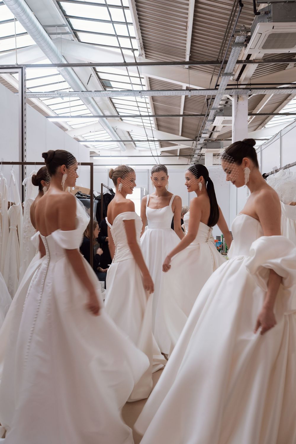 Five models in white bridal gowns stand in a circle in a bridal showroom with racks of wedding dresses.