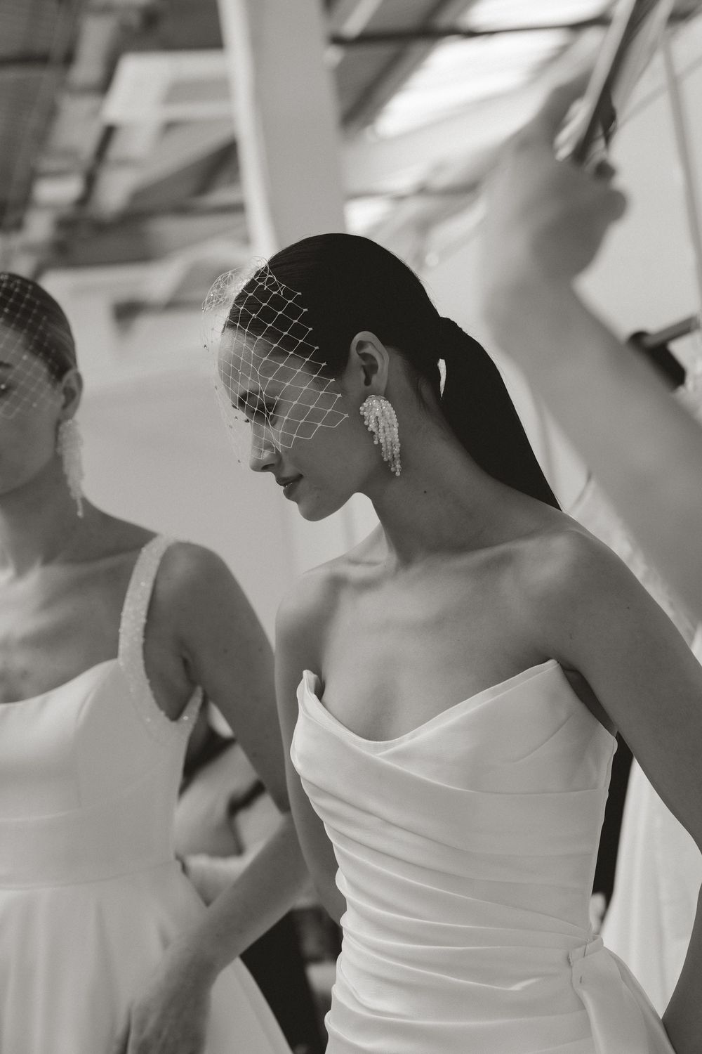 Black and white photo of a bride in a strapless white gown wearing a birdcage veil and pearl earrings.