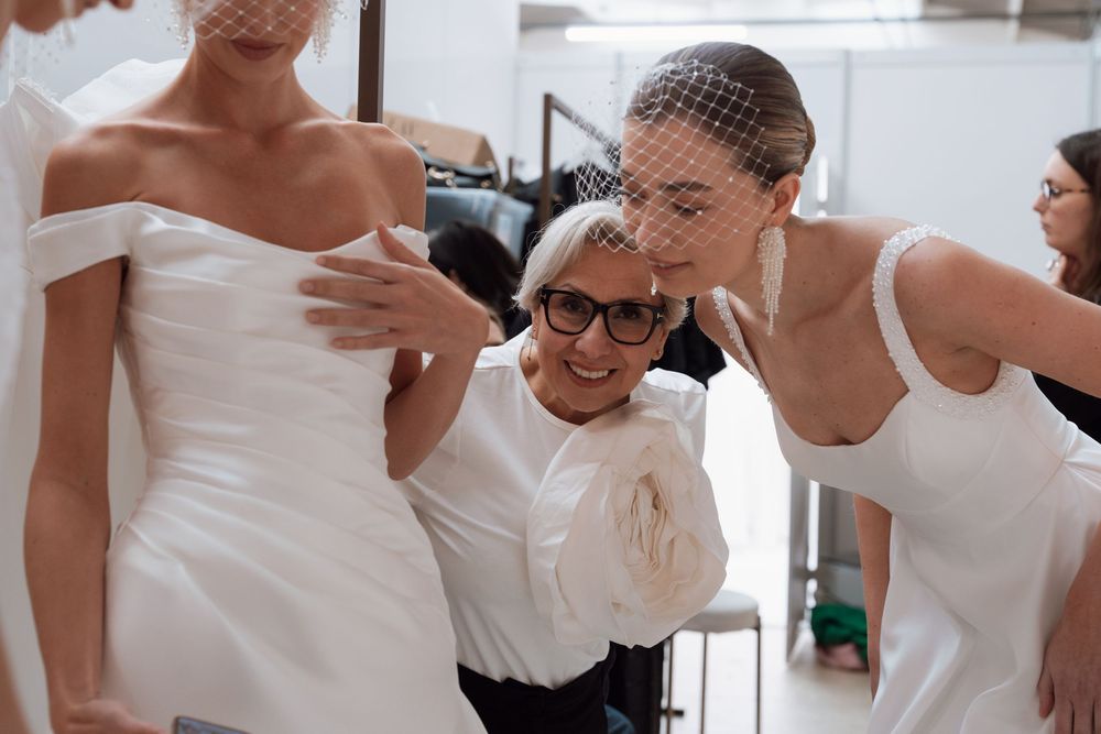 Fashion designer adjusting white bridal gowns on models backstage at a wedding dress fitting session.