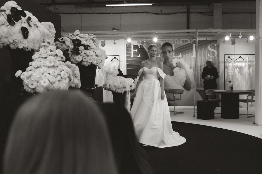 Black and white photo of a bride in a ballgown at a bridal expo, surrounded by floral arrangements and wedding dress displays.
