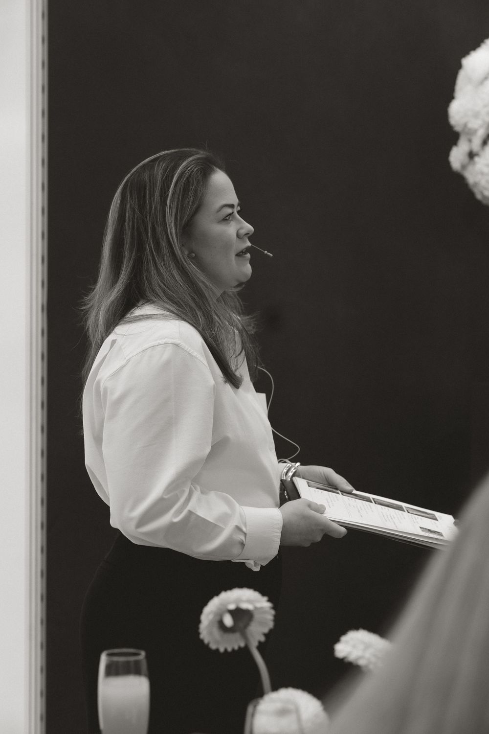 Black and white photo of a woman in a white blouse holding a book, speaking at a professional event.