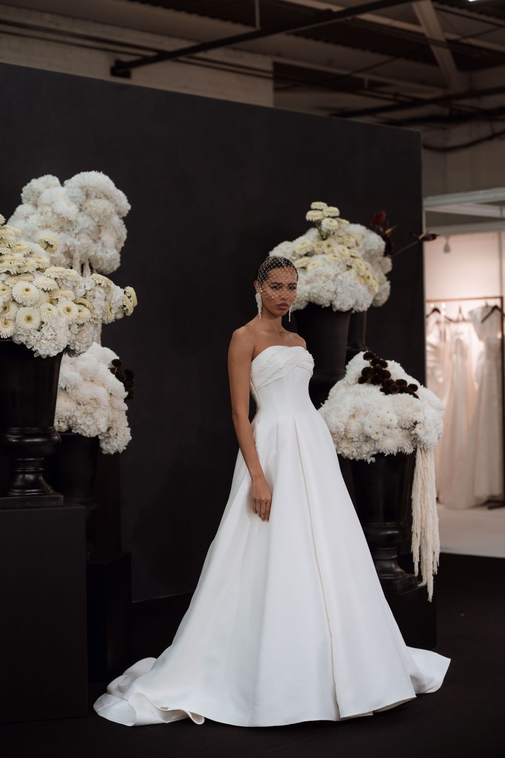Model wearing a strapless white ball gown wedding dress, posed between large white floral arrangements on black pedestals.