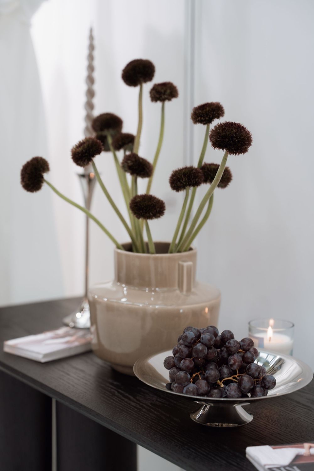Dark dried flowers in beige ceramic vase beside silver cake stand with grapes and lit candle on dark wood shelf.