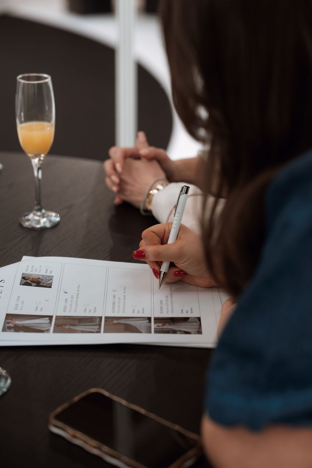 Woman writing on a printed document at a dark table with a champagne mimosa glass and smartphone nearby.
