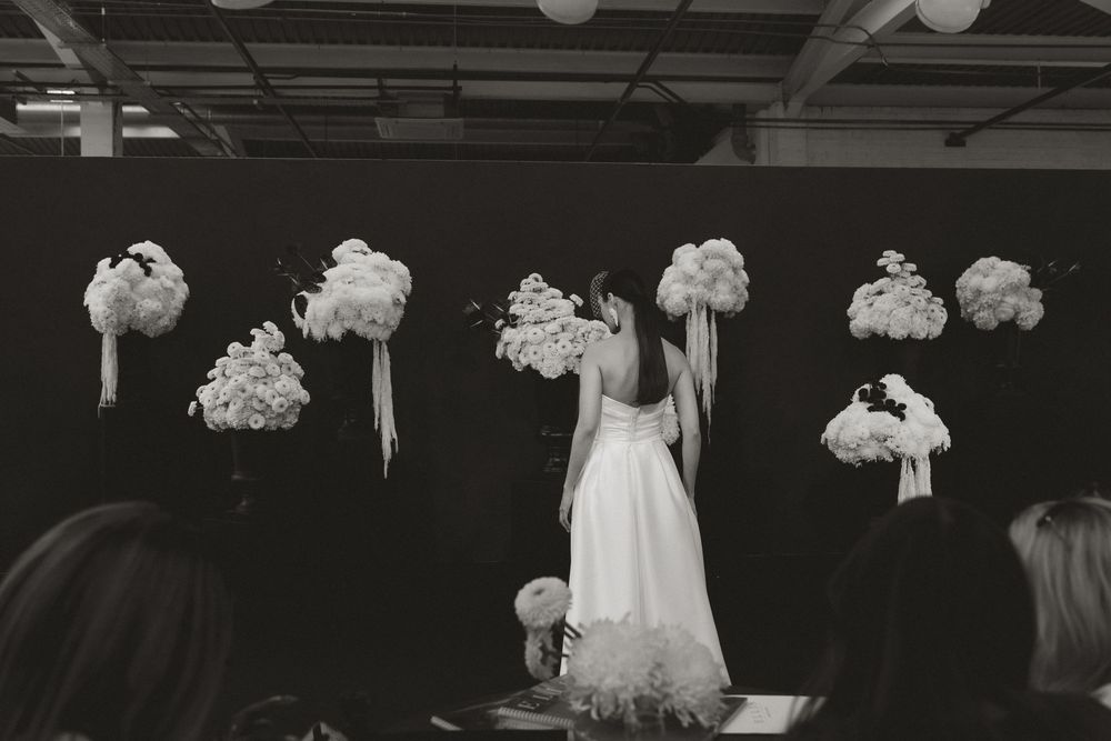 Black and white photo of a bride standing among elegant floral arrangements with white bouquets at a wedding ceremony.