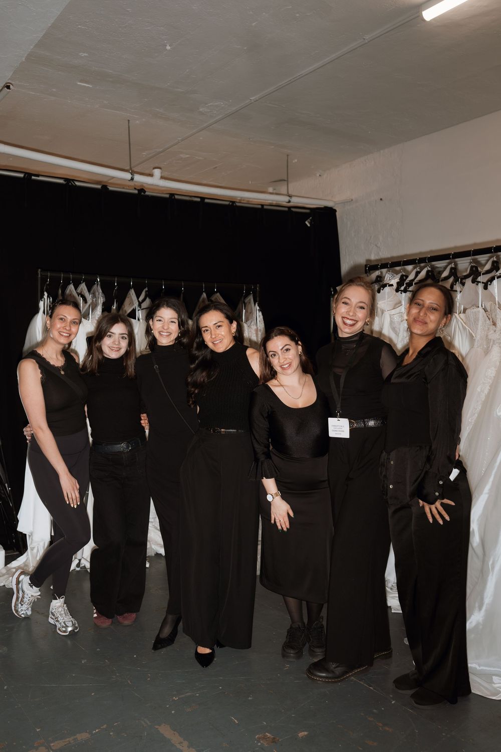 Seven women dressed in black pose together in a bridal boutique backstage area with white gowns on racks.