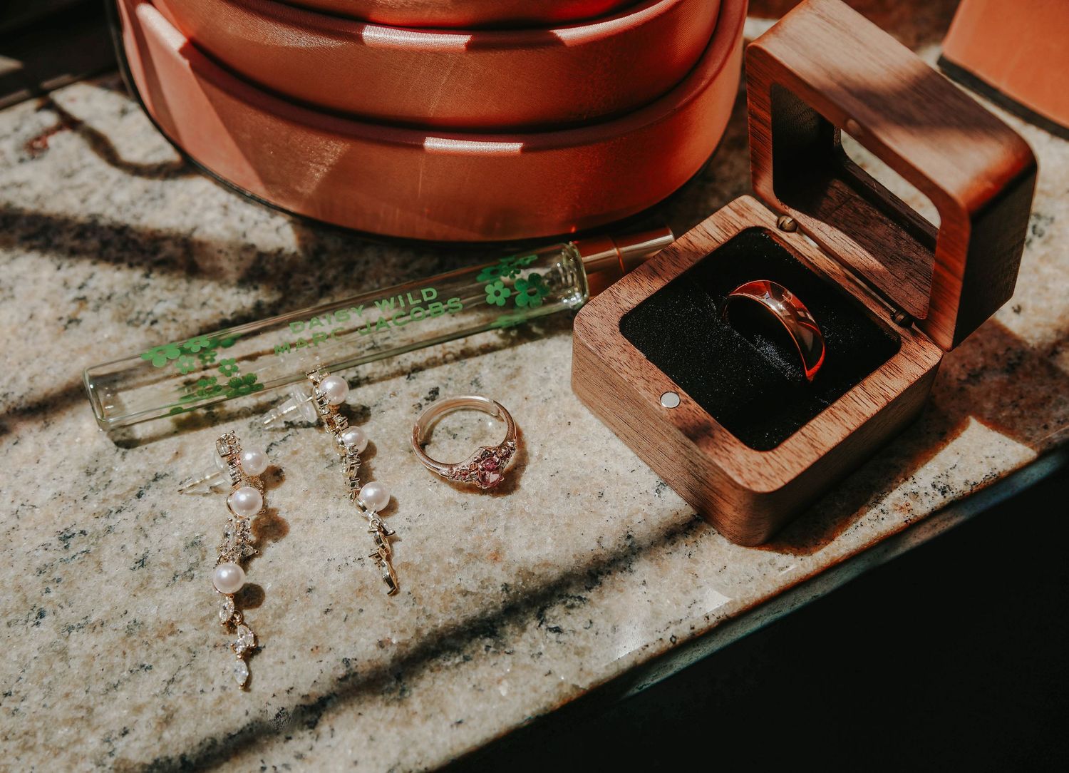 Wedding rings in wooden box beside diamond earrings, gold ring, and green pen on marble surface with leather belt.