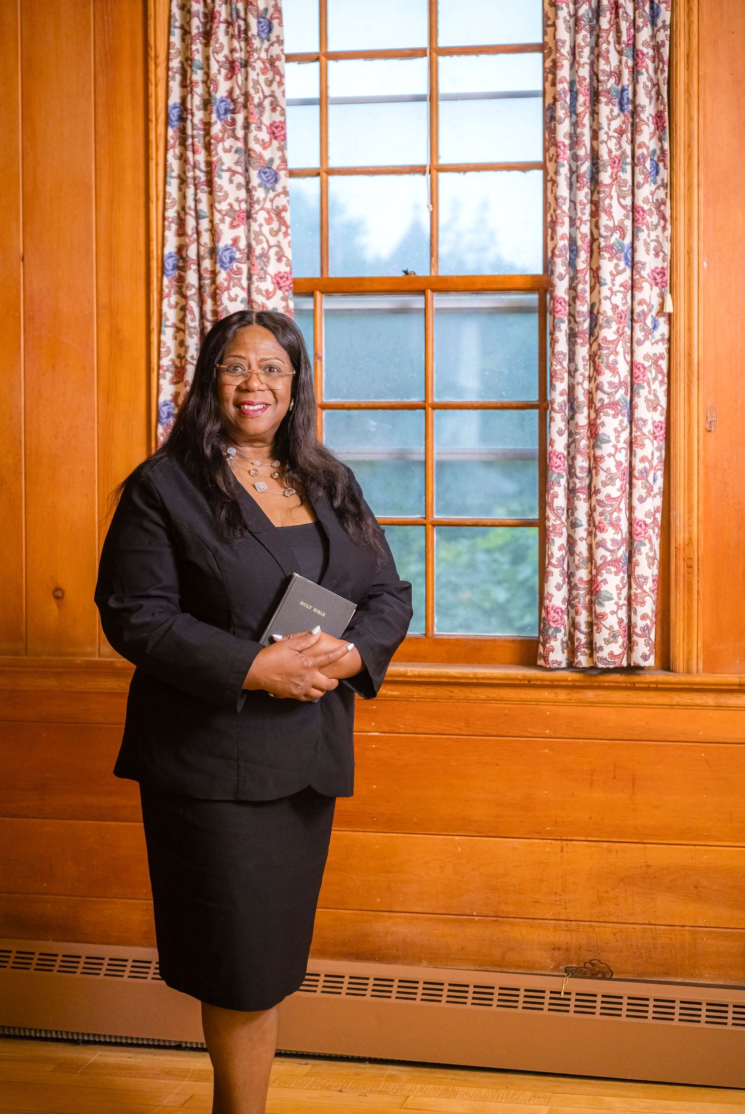 Professional woman in black suit holding a book, standing by a window with floral curtains in a wood-paneled room.