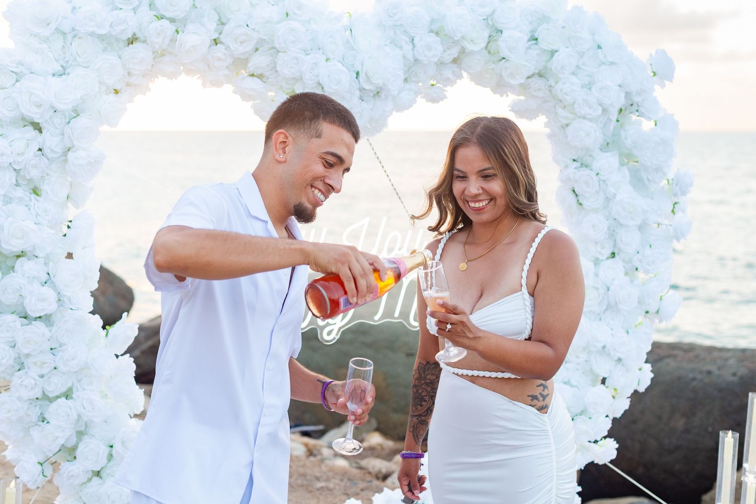 Marriage proposal. Toast. Man, woman, and decorative setup with white flower heart overlooking the ocean in Río Grande, Puerto Rico.