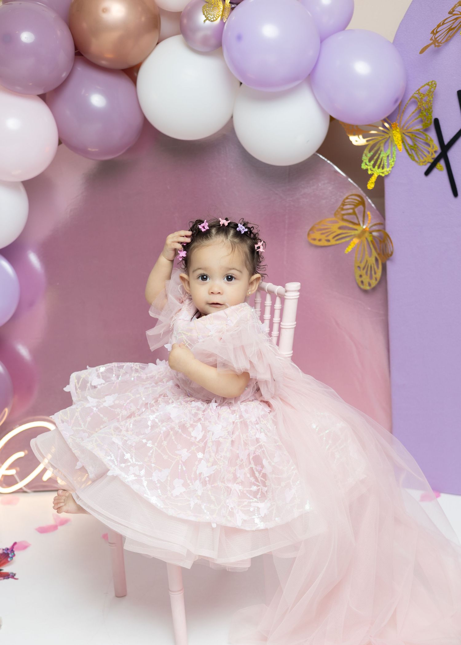 A toddler in a pink tulle dress sits on a chair surrounded by purple and pink balloons and gold butterfly decorations.