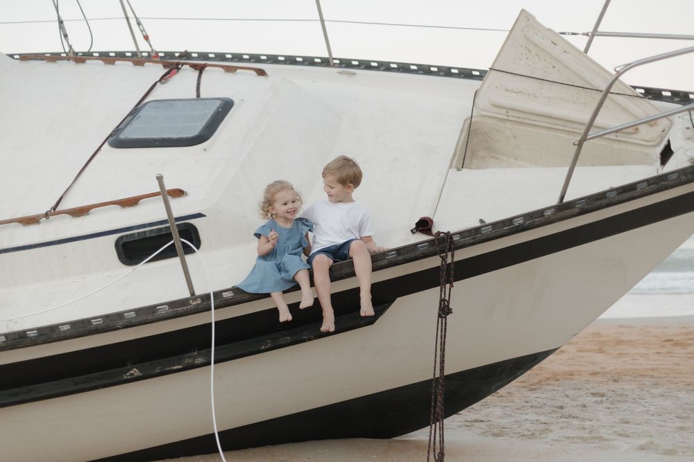 Two young children sitting on the bow of a beached sailboat on a sandy shore.