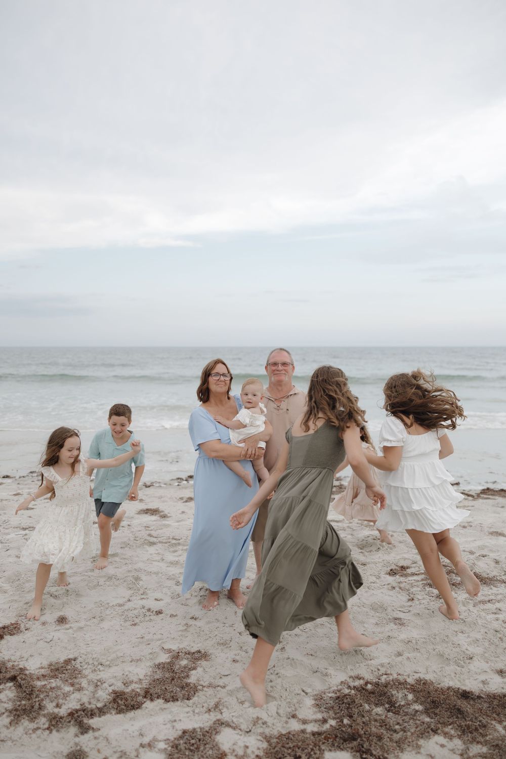 Multi-generational family playing on a sandy beach at dusk with children running toward grandparents holding a baby.