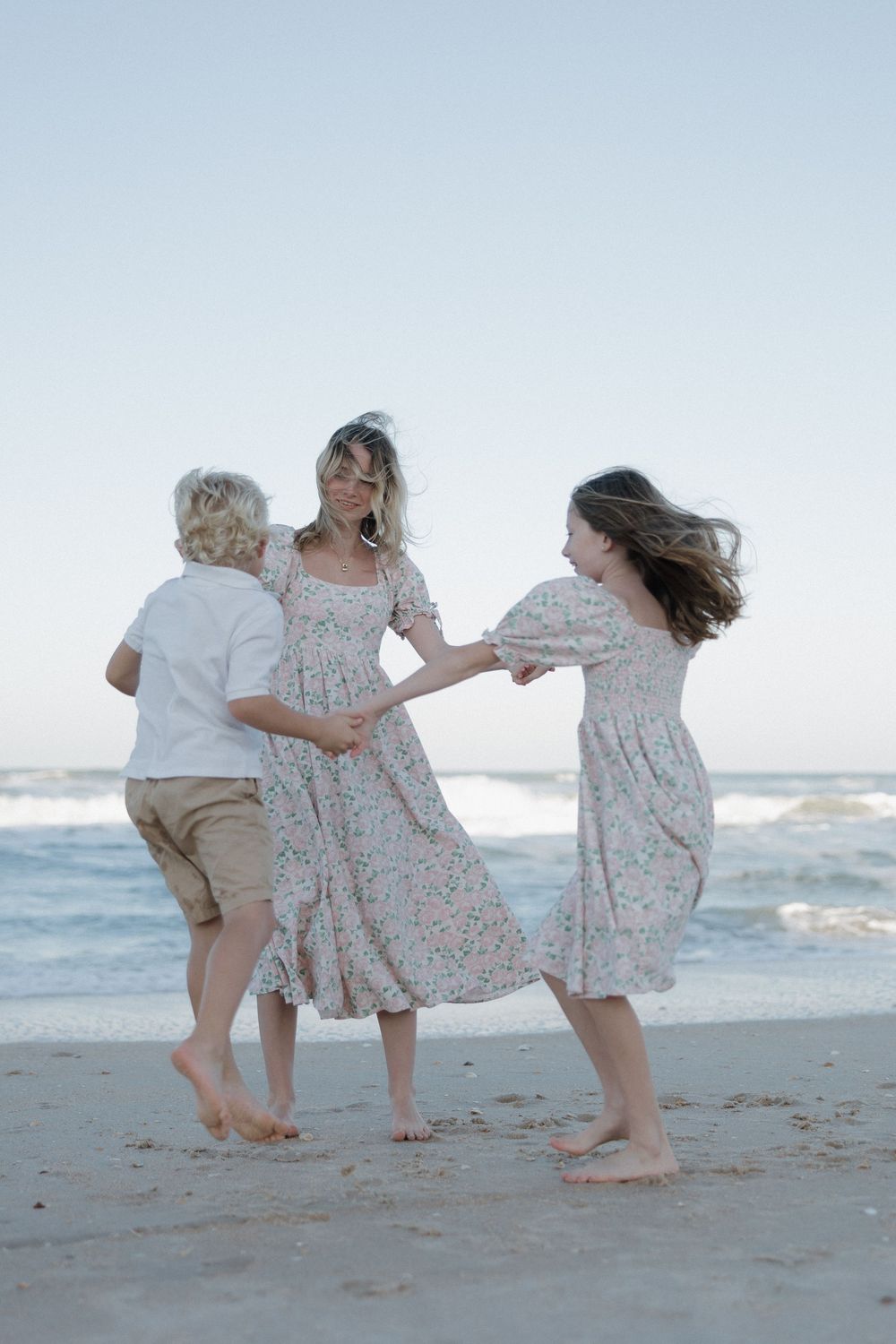 Mother and two children dance joyfully barefoot on a sandy beach at dusk near ocean waves.