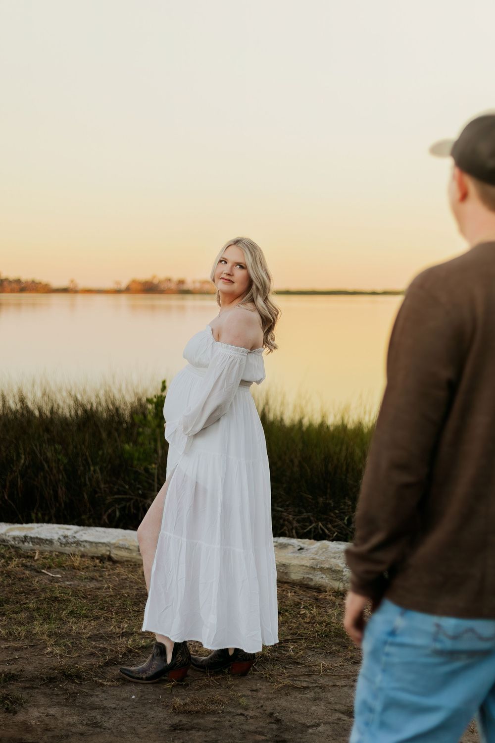 Blonde woman in white off-shoulder maxi dress smiles over shoulder at sunset by a lake with a man in foreground.