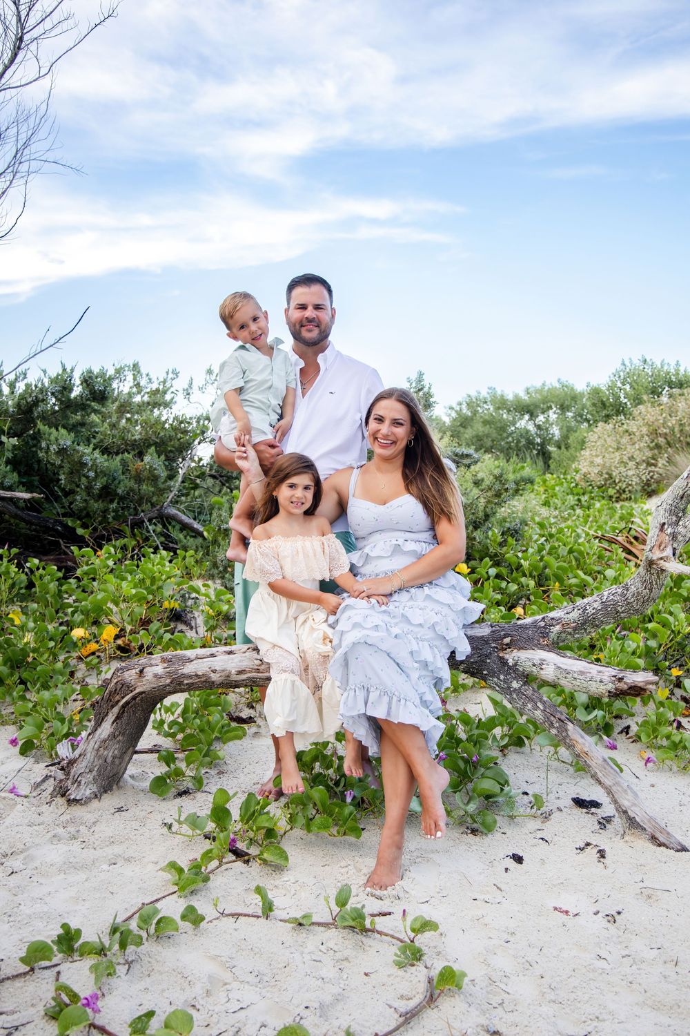Family of four poses on driftwood at a sandy beach, surrounded by lush greenery under a cloudy sky.