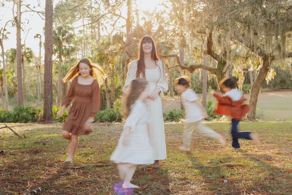 Woman in white dress stands outdoors while children play around her in a sunlit park with moss-draped trees.