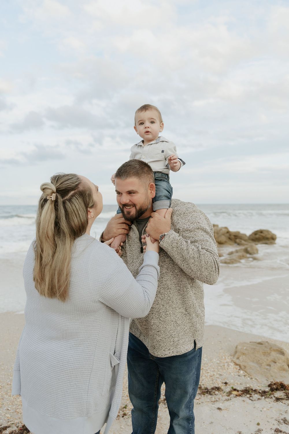 Young couple with baby on shoulders enjoys a candid moment on a rocky ocean beach on a cloudy day.