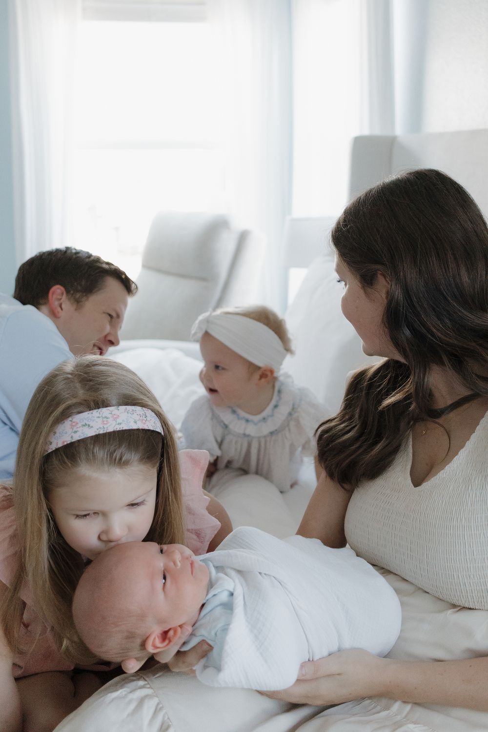Family with newborn baby, two young girls, and parents sharing a tender moment on a white bed.