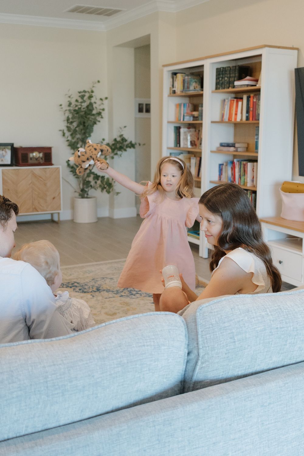A young girl in a pink dress holds flowers while playing with family in a bright, cozy living room.