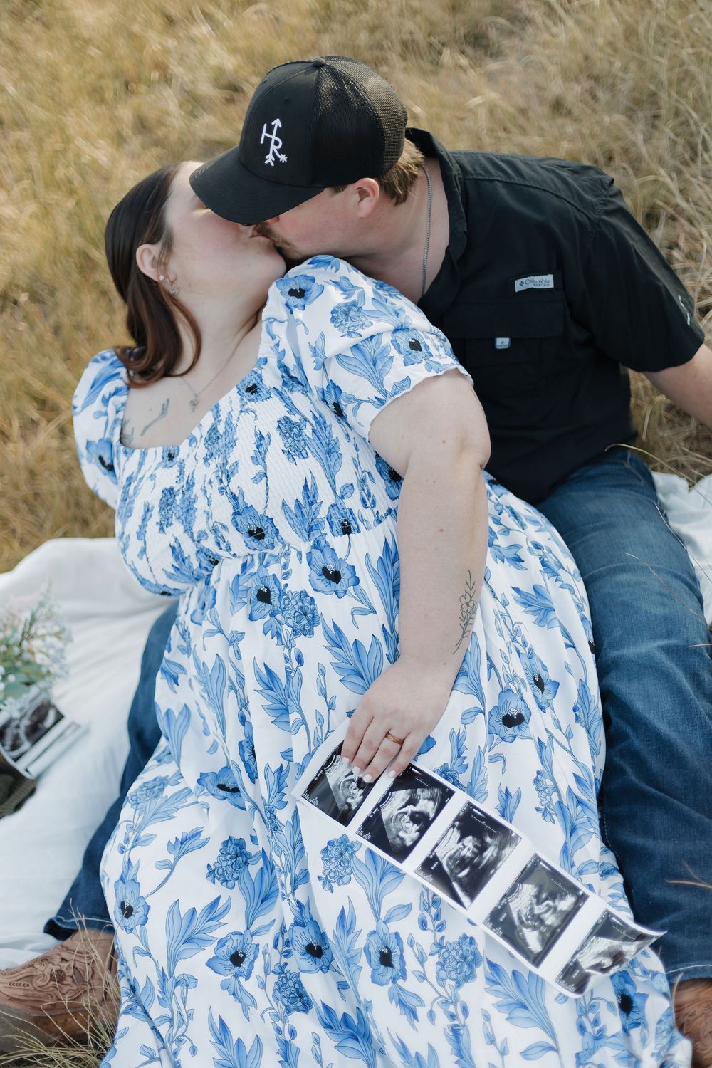 Couple kissing on a blanket in a field holding ultrasound photos, woman wearing a blue floral maternity dress.