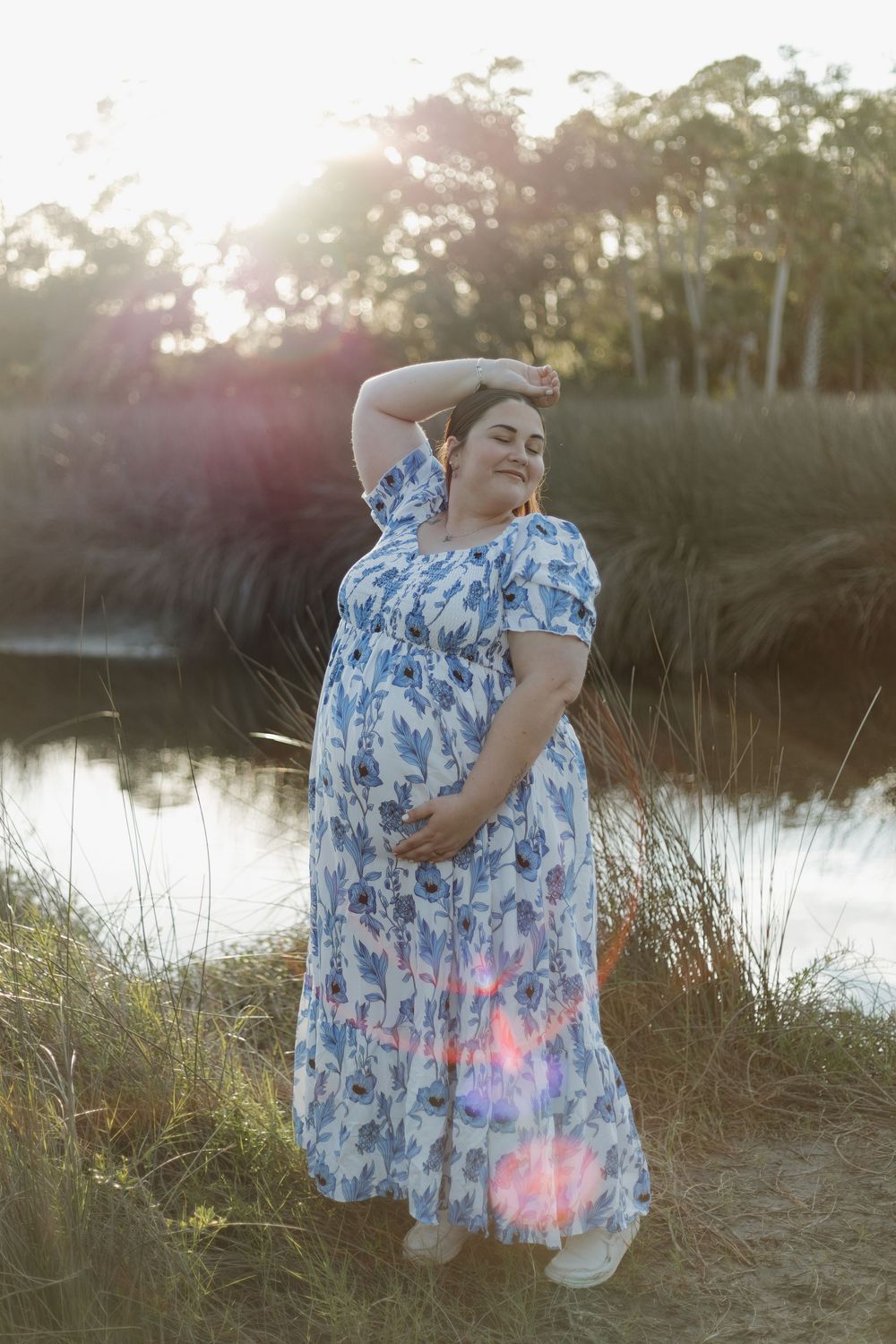 Pregnant woman in floral maxi dress poses gracefully by a sunlit pond surrounded by tall grass.
