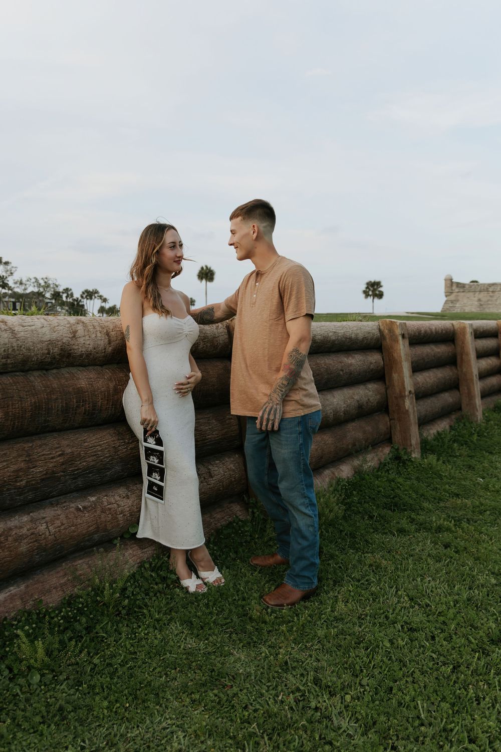 Pregnant woman in white dress holds ultrasound photos while partner places hand on her shoulder near rustic log wall outdoors.