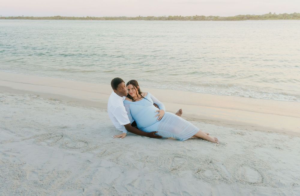 Couple sitting on white sand beach at sunset, pregnant woman in blue dress leaning against partner near calm water.
