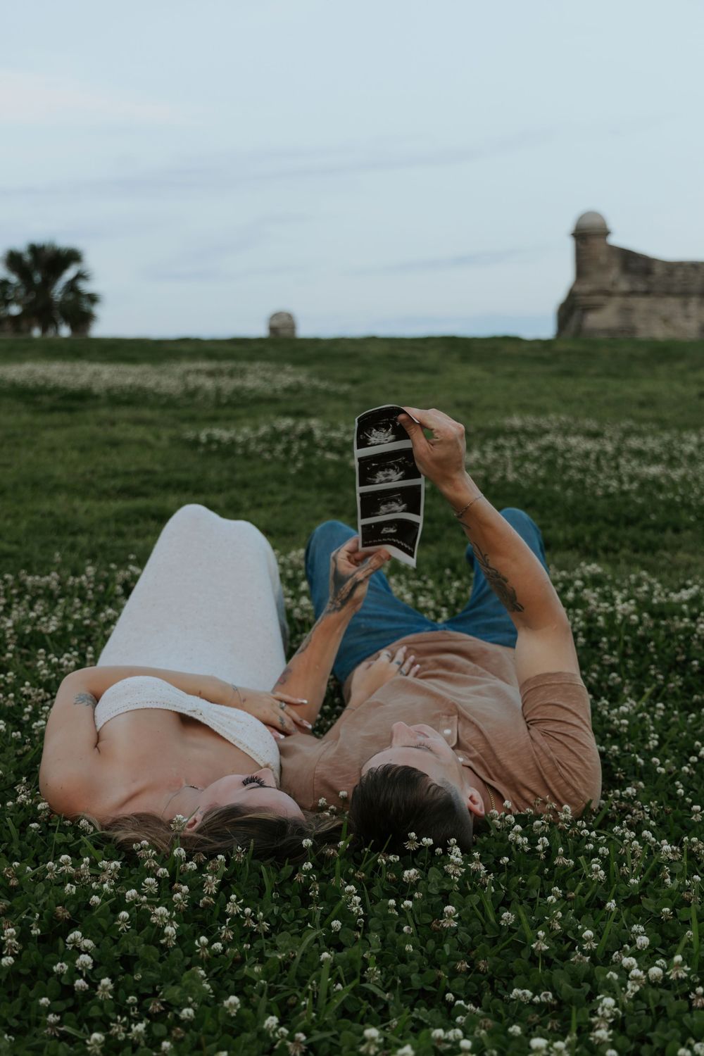 Couple lying in a flower field holding ultrasound photos, announcing pregnancy near a historic fort.