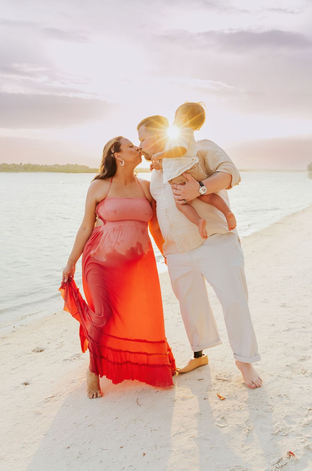 Pregnant woman in red dress with partner holding toddler sharing a kiss on a sunny beach at sunset.