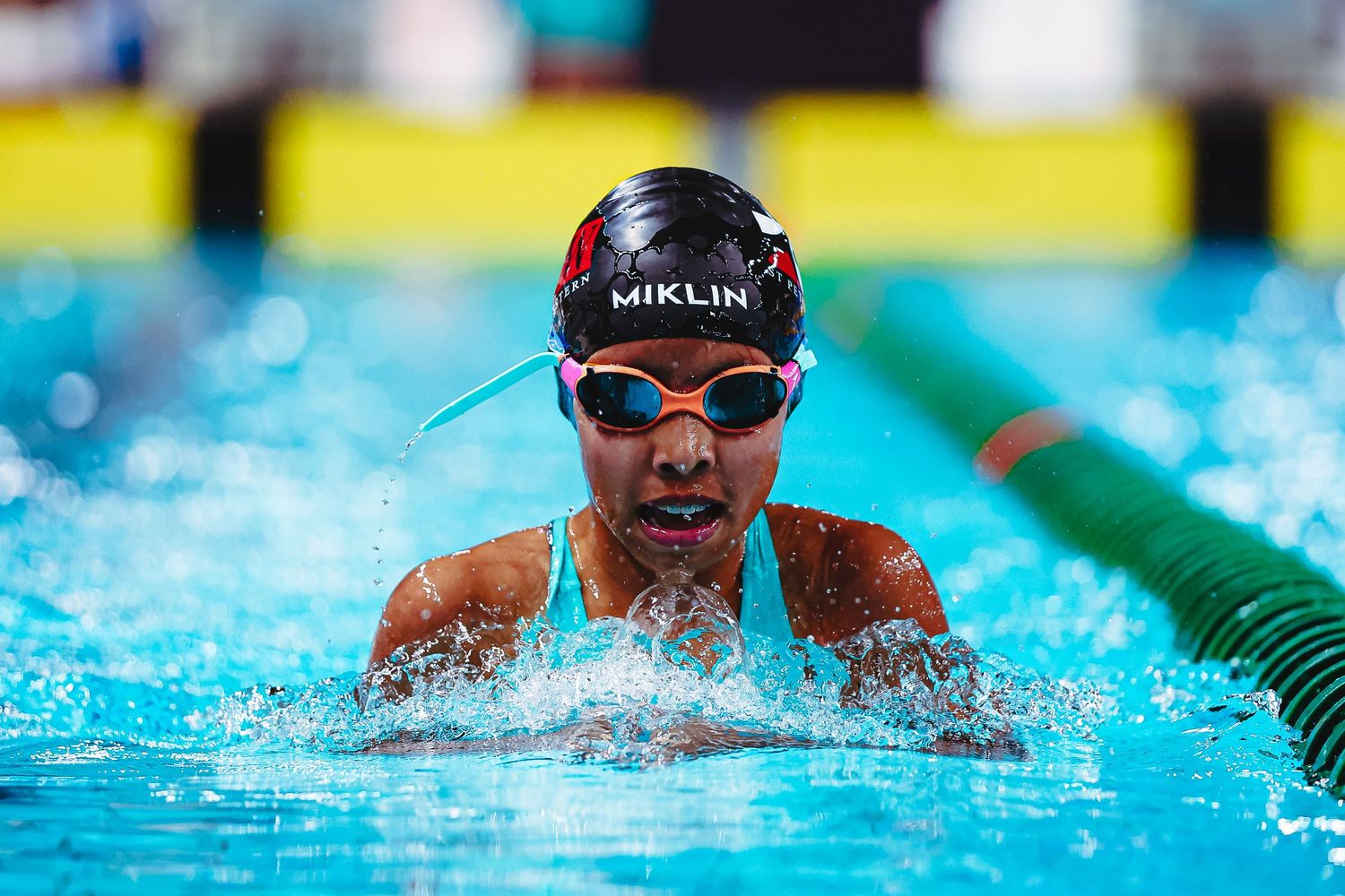 Young female swimmer smiling poolside wearing a Speedo black swimsuit and colorful orange swim cap at an indoor competition.