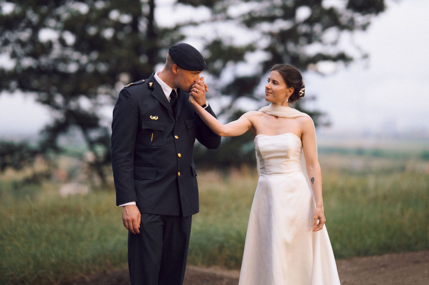 Military groom in uniform kisses bride's hand outdoors on their wedding day.