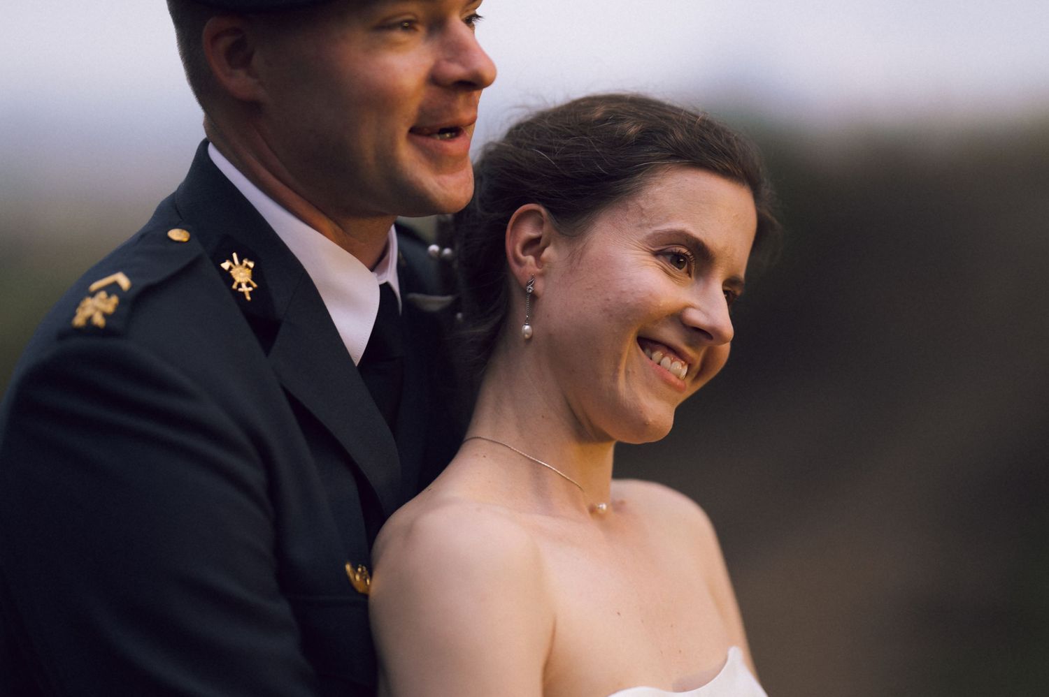 A smiling bride in a white strapless gown stands with a groom in military dress uniform outdoors.