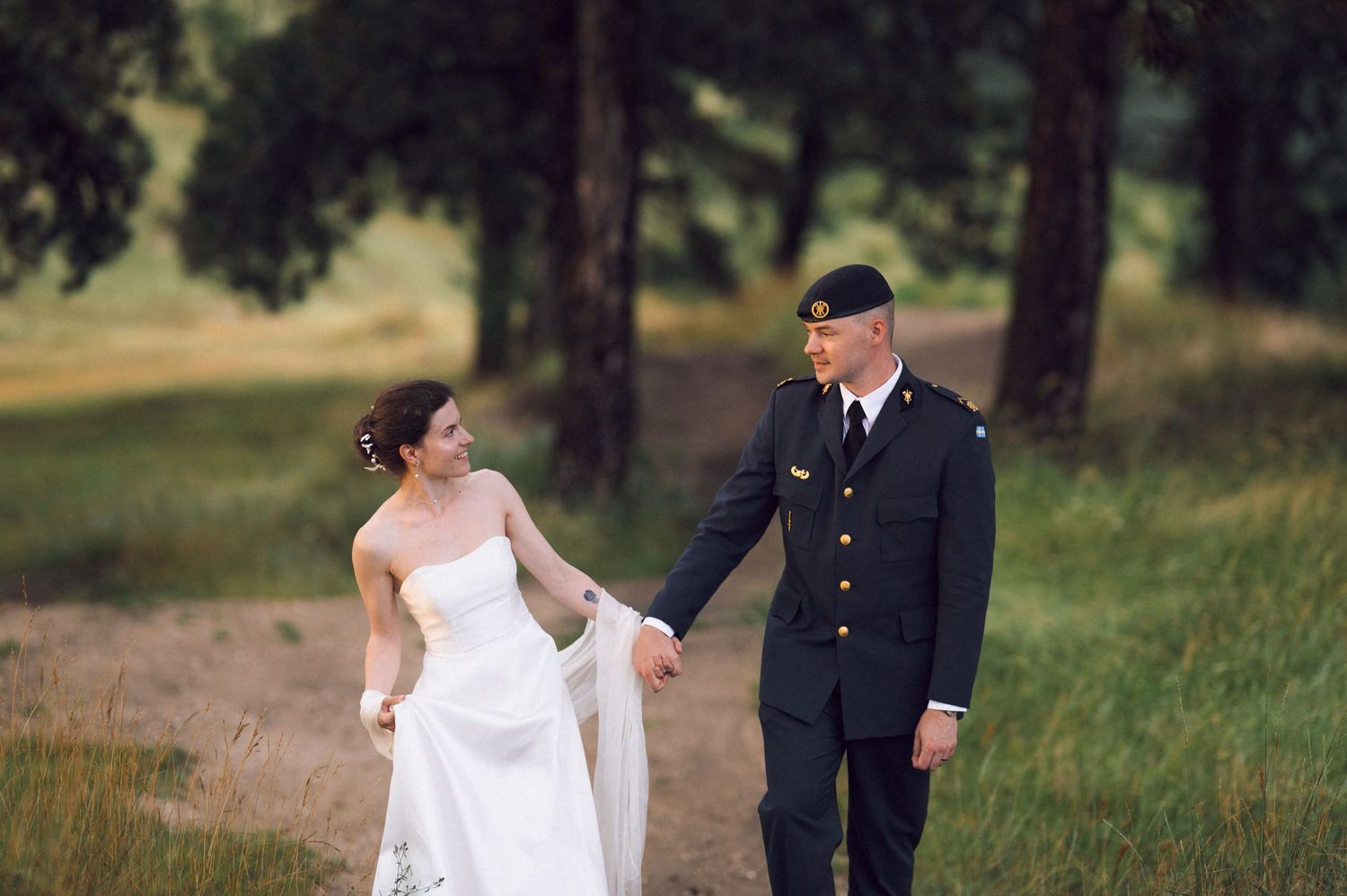 Military groom in uniform holds hands with bride in white dress walking on a scenic outdoor path surrounded by trees.