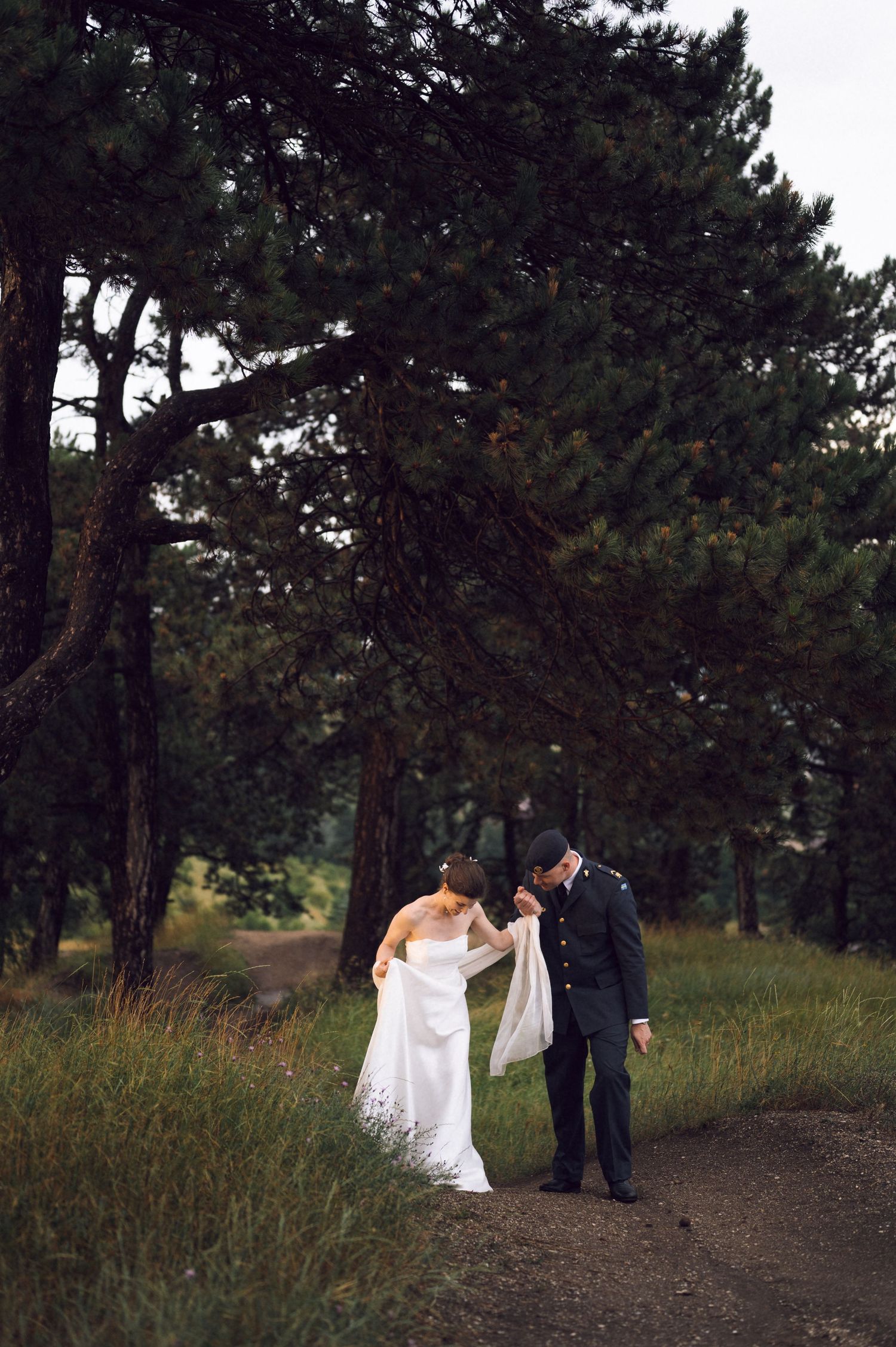 A bride in a white gown and groom in dark military suit walk hand in hand along a forest path surrounded by tall pine trees.