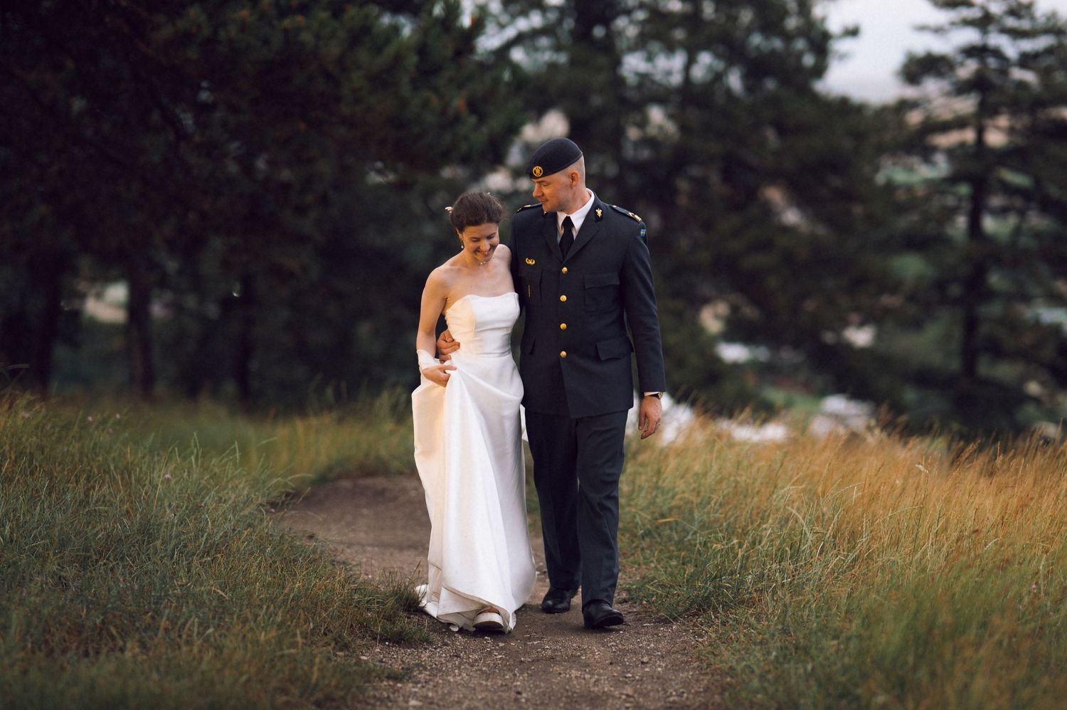 A military groom in dress uniform walks with his bride in a white gown along a scenic path surrounded by tall grass and trees.