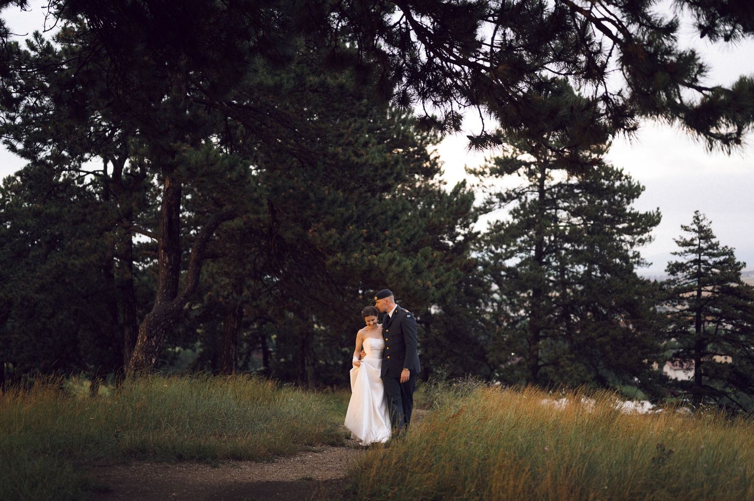 Bride and groom walking on a forest path surrounded by tall pine trees and golden grass at dusk.