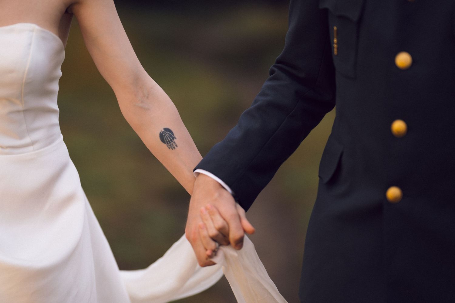 Bride and groom holding hands, showing tattoo on wrist, military uniform with gold buttons visible.