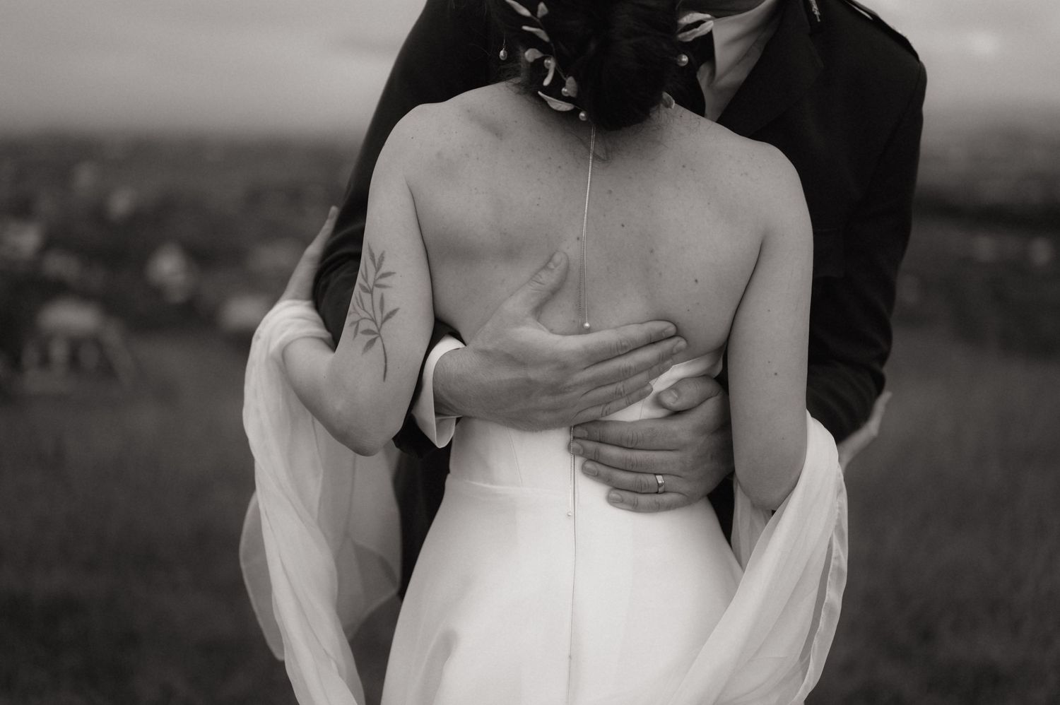 Black and white wedding photo of groom embracing bride from behind, showing her open back dress and floral tattoo.