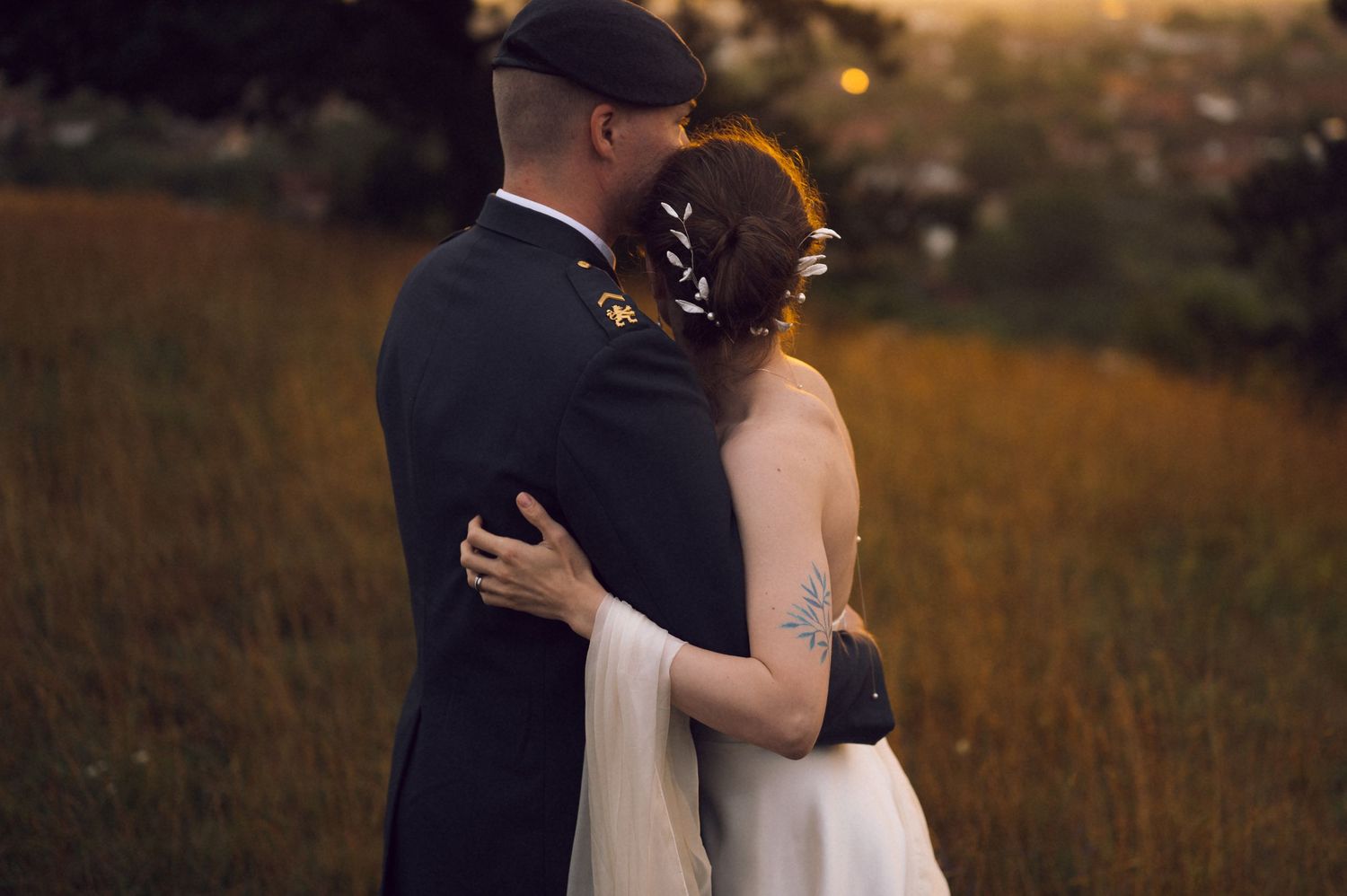 Military groom and bride in white dress embrace at golden hour in a field, bride showing a blue tattoo on her arm.
