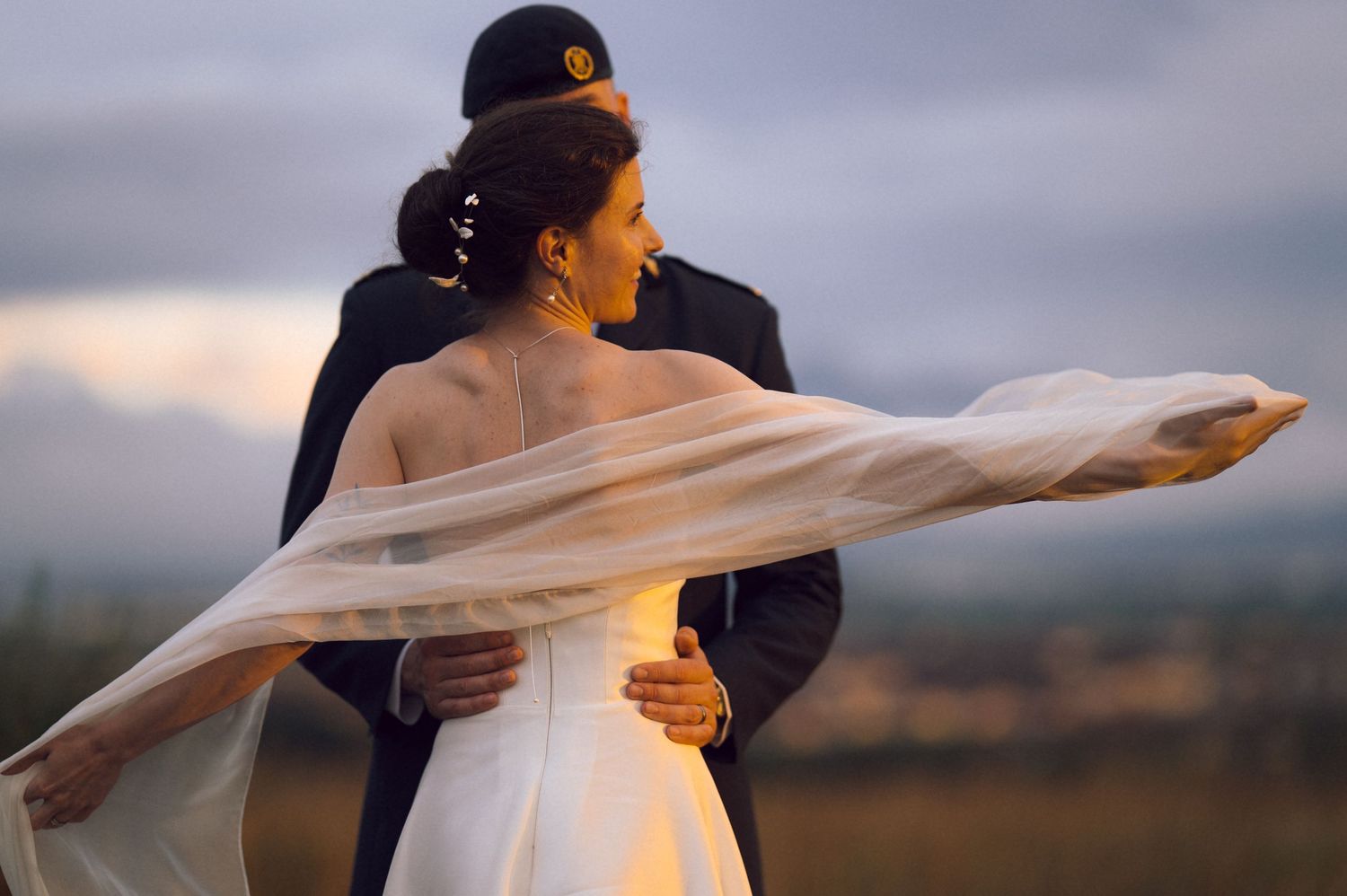 Bride in white gown with flowing veil embraced by groom in dark suit during golden hour outdoor wedding photo.