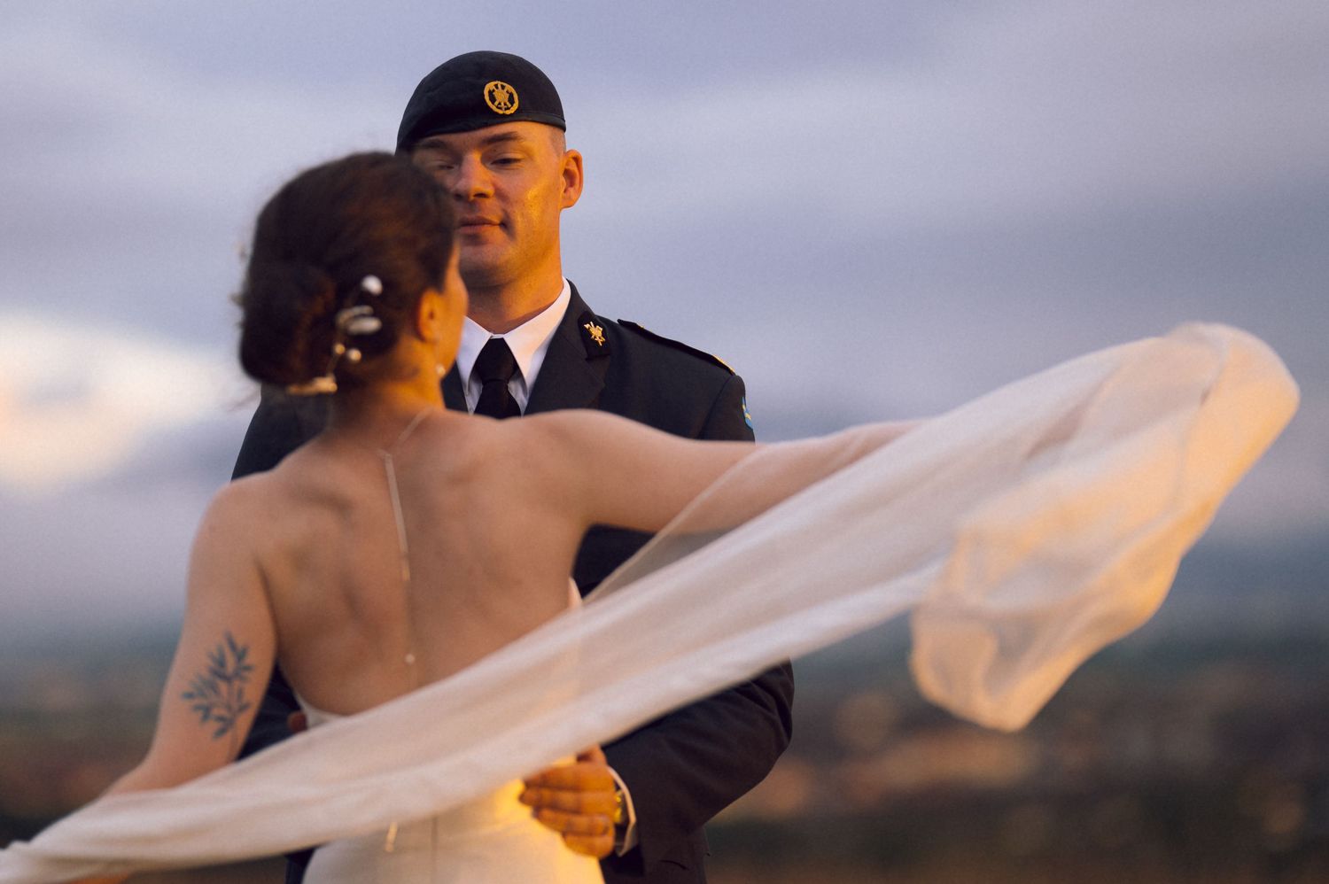 Military groom in uniform embraces bride in backless wedding dress, her veil flowing in the wind against a scenic sky.