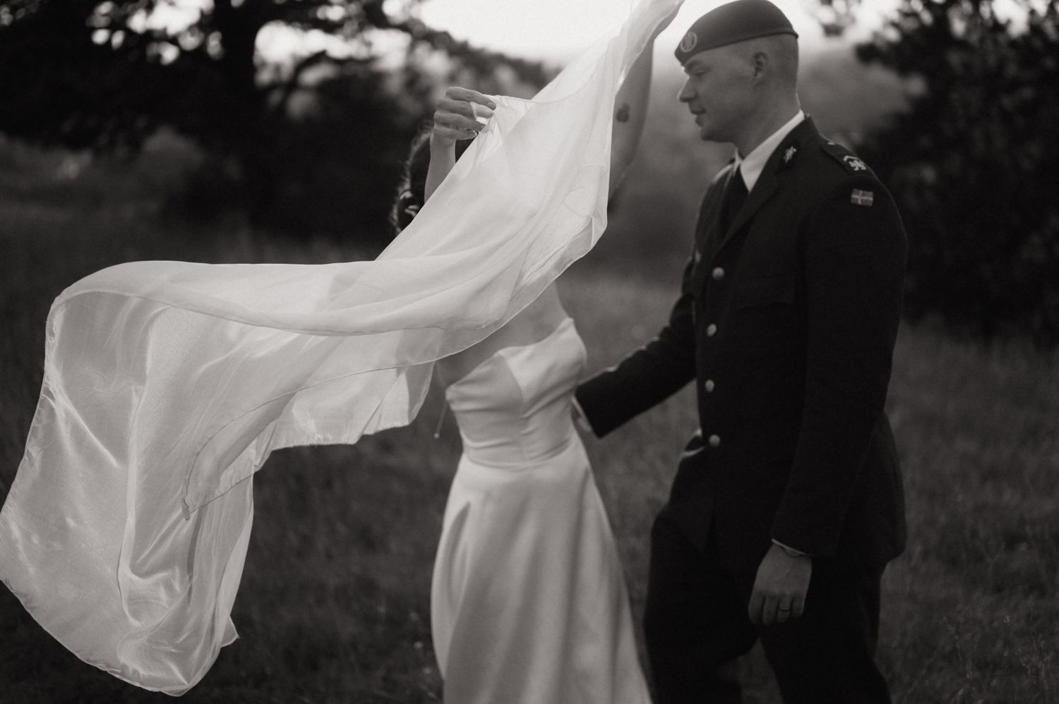 Black and white photo of a military groom and bride, her veil blowing in the wind during an outdoor wedding.
